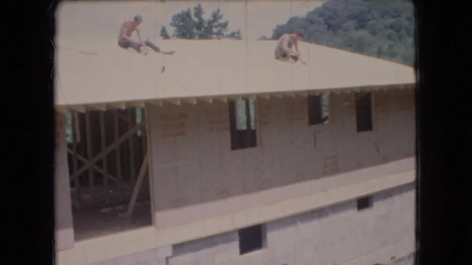 Ohio 1966 home-movie frame of men working or seated on the roof of a partially built house, with wall sheathing, open structural framing, a concrete block foundation, and trees in the background.