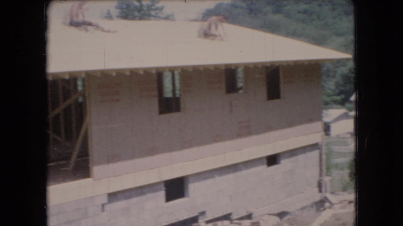 1966 Ohio 8mm frame showing two men on the roof of a two-story house under construction, with sheathing, foundation, trees, and distant homes visible.