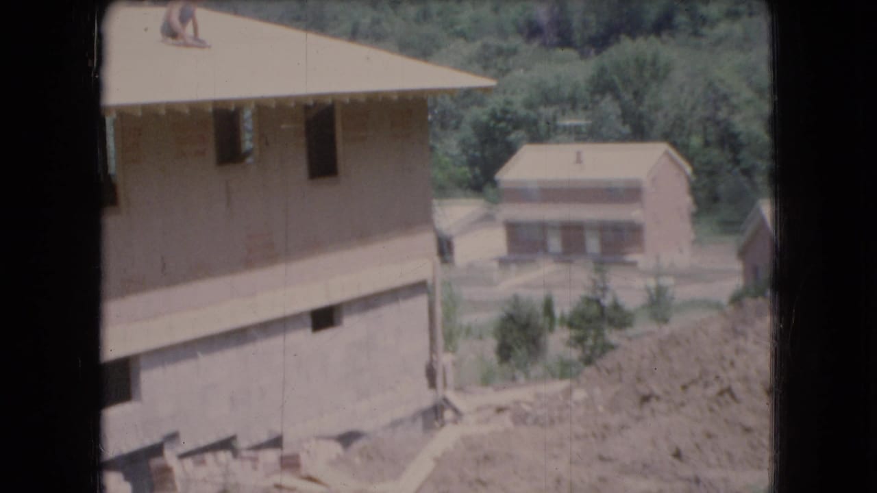 A man sits on the roof of an unfinished house while another house appears in the background.