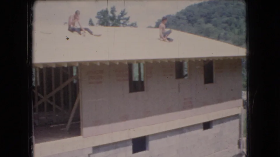 1966 Ohio 8mm frame showing two men on a pale sloped roof over a partially framed house, with open wall framing, foundation blocks, and trees in the background.