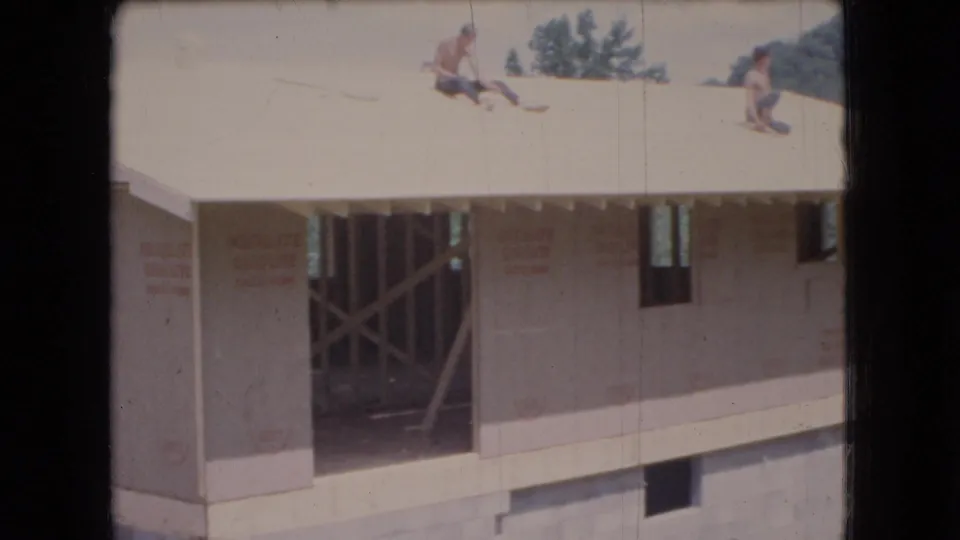 1966 Ohio home-movie frame of two men on the roof of a house under construction, showing exposed supports, wall sheathing, a foundation opening, and wooded surroundings.