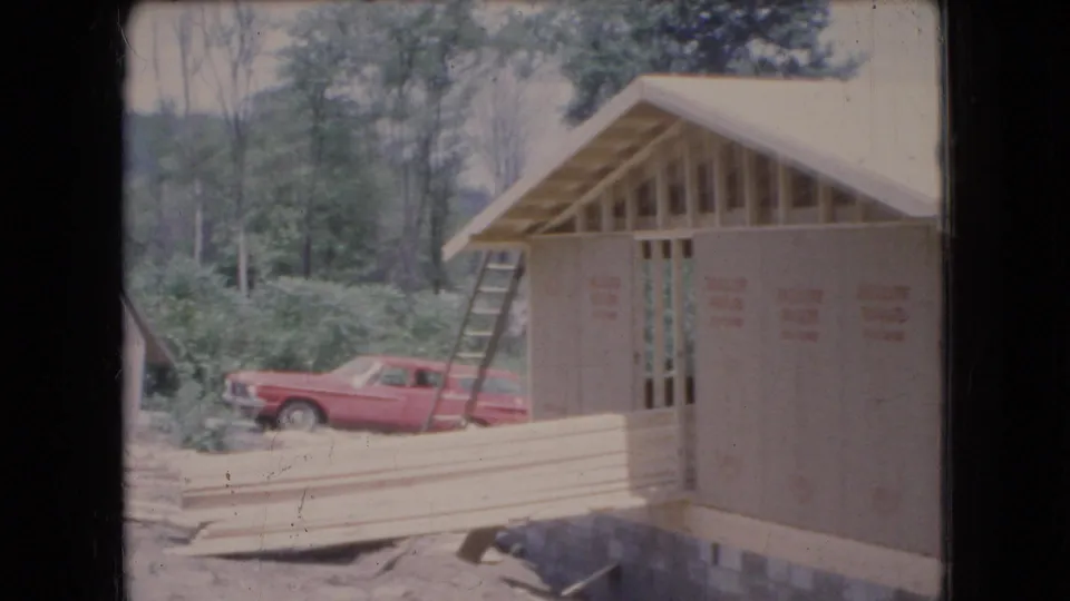 1966 Ohio frame showing a garage under construction beside stacked lumber, a ladder, wooded brush, and a red 1960s station wagon parked nearby.
