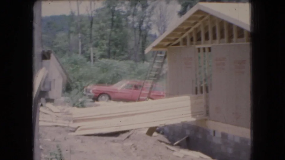 1966 Ohio 8mm frame of the garage build from another angle, with a ladder against the structure, stacked boards in front, and a red station wagon near the trees.