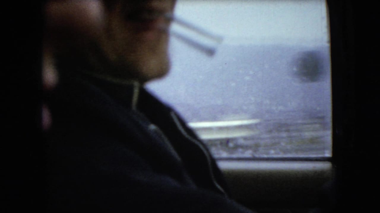 Adult male driver in profile, cigarette held at his mouth, seen inside a dark car with rain-softened roadside detail beyond the window in the supplied 1966 Ohio footage.