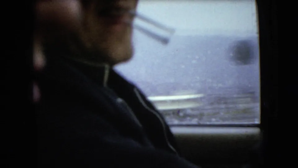 Adult male driver in profile, cigarette held at his mouth, seen inside a dark car with rain-softened roadside detail beyond the window in the supplied 1966 Ohio footage.