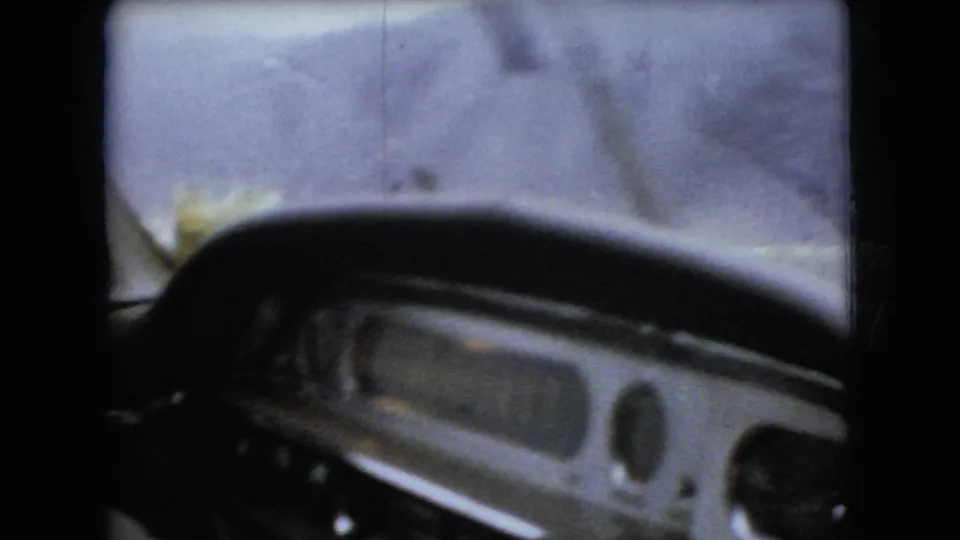 Passenger-seat view of the steering wheel, dashboard, windshield, and wiper blur as the car moves through rainy conditions in the supplied 1966 Ohio home-movie clip.