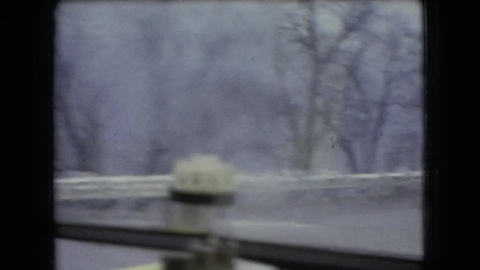 Rain-softened windshield view from inside the car showing a wet roadside guardrail, blurred trees, and dashboard detail in the supplied 1966 Ohio 8mm footage.