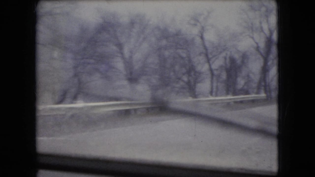 1966 Ohio 8mm frame from a moving vehicle showing a wet roadside curve, guardrail, bare trees, and dark window edges under an overcast sky.