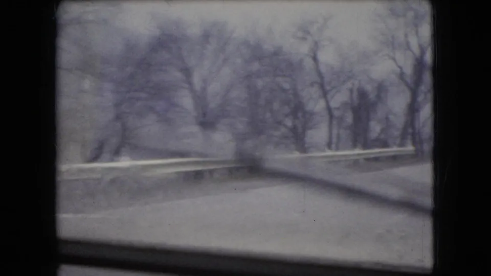 1966 Ohio 8mm frame from a moving vehicle showing a wet roadside curve, guardrail, bare trees, and dark window edges under an overcast sky.