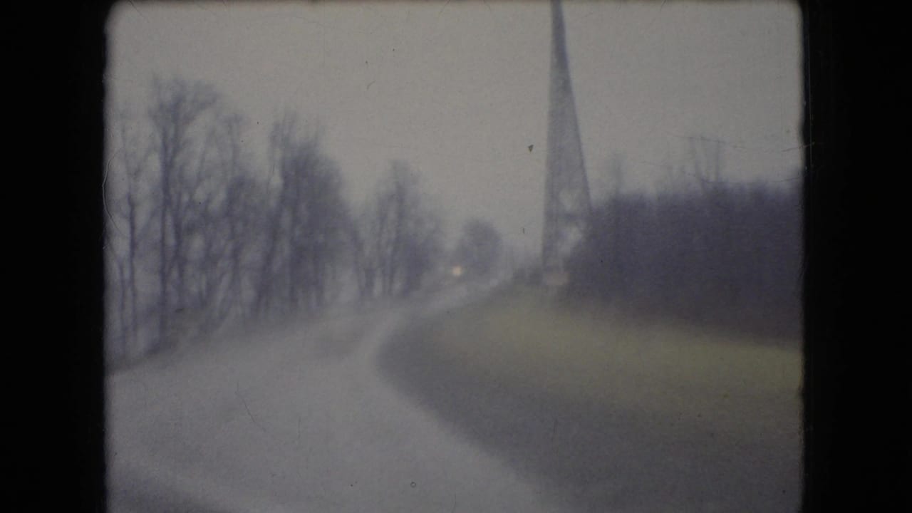 Curving wet road with bare trees and a tall tower-like structure in the distance.