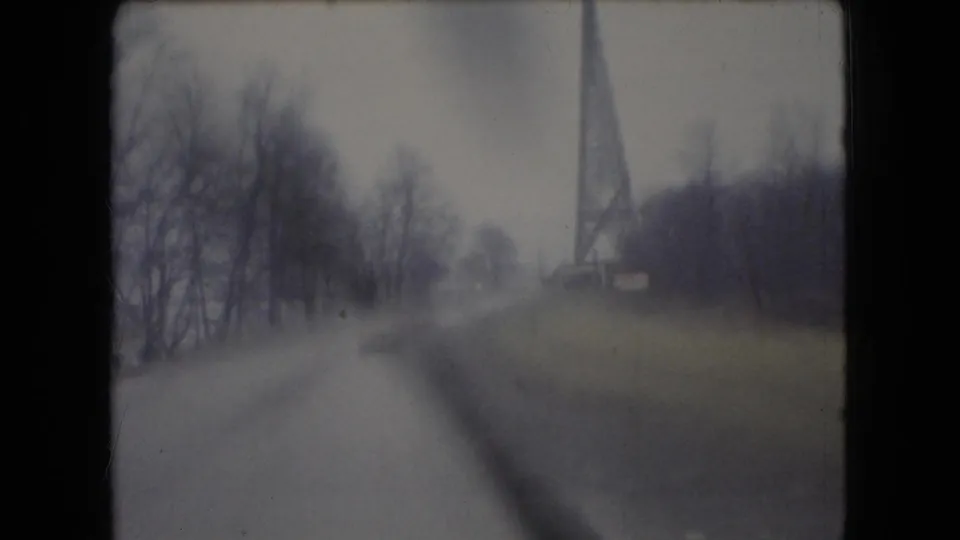 1966 Ohio 8mm frame from inside a moving car, with a wet road, leafless trees, window edge, and distant tower-like structure softened by motion blur.