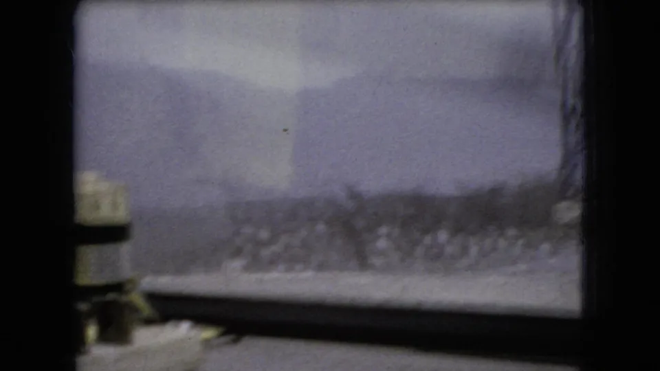 1966 Ohio home-movie frame from the car interior, with a small dashboard-mounted object, divided windshield, cloudy sky, and passing rural hillside visible beyond the glass.