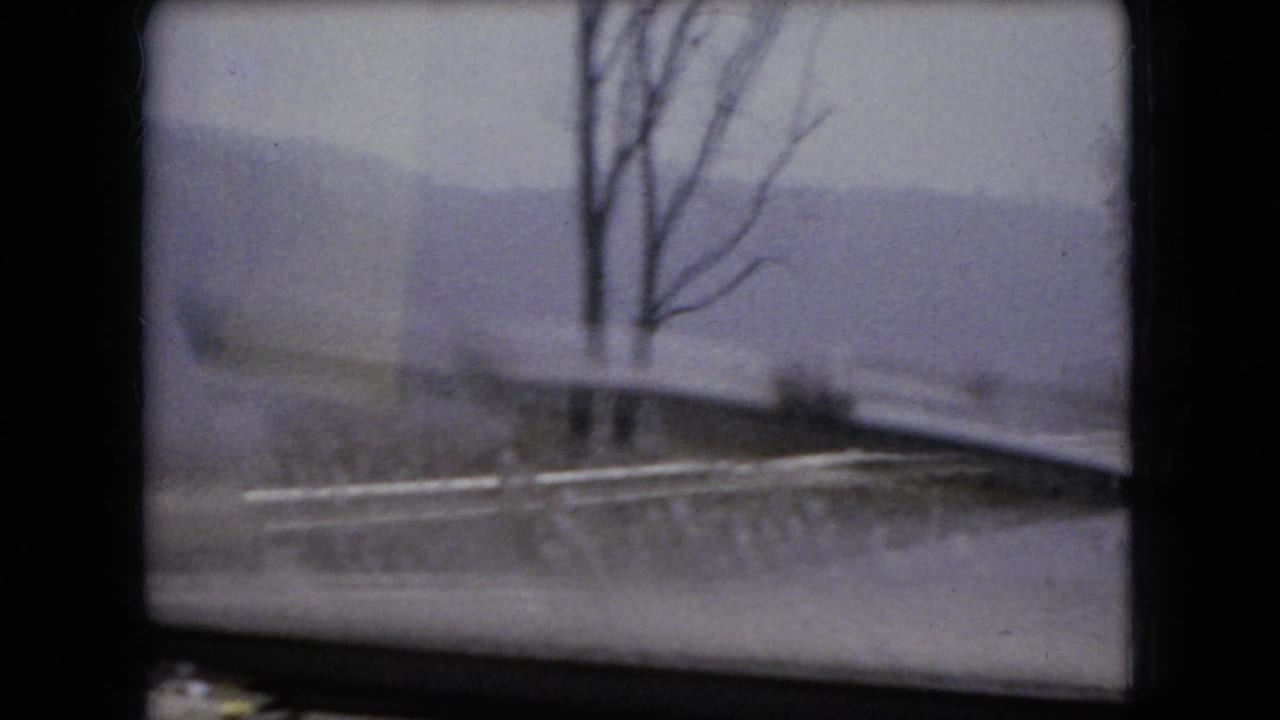 1966 Ohio home-movie frame looking through a car windshield at a snowy roadside, bare trees, and a muted winter landscape beyond the glass.