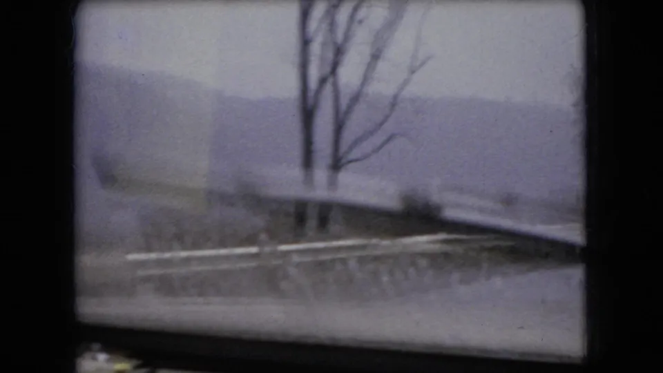 1966 Ohio home-movie frame looking through a car windshield at a snowy roadside, bare trees, and a muted winter landscape beyond the glass.
