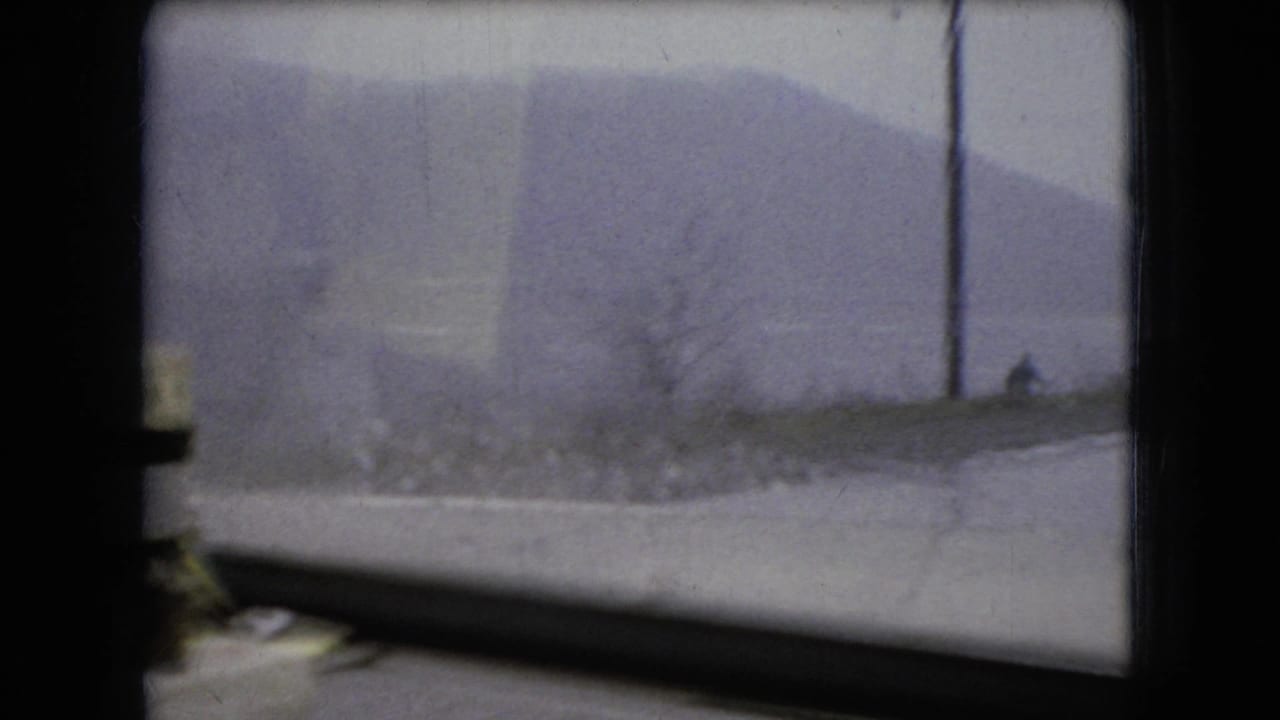 1966 Ohio home-movie frame looking through wet divided car glass toward a gray roadside landscape and distant slope.