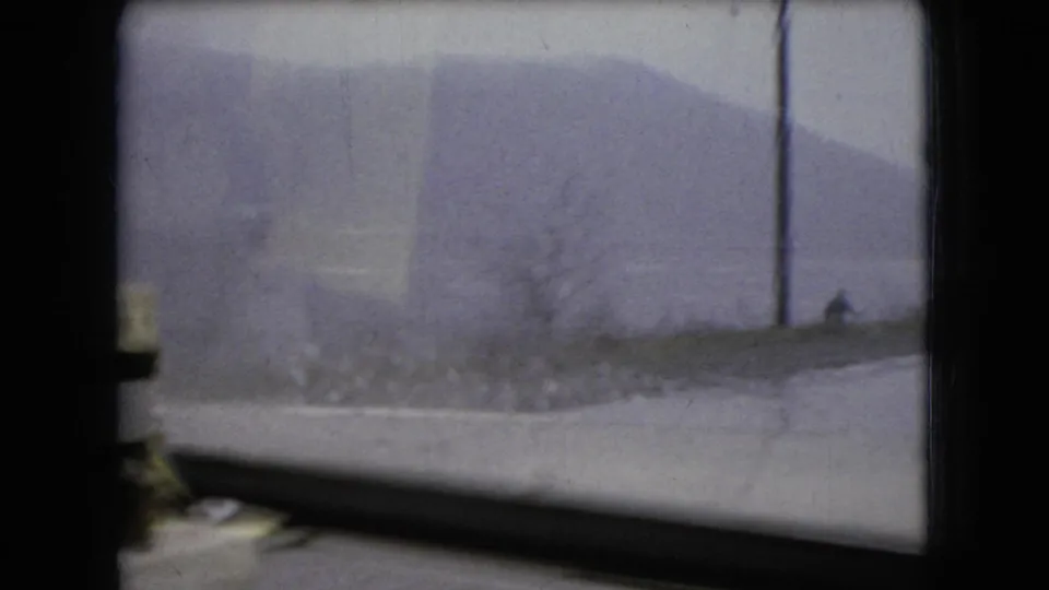 1966 Ohio home-movie frame looking through wet divided car glass toward a gray roadside landscape and distant slope.