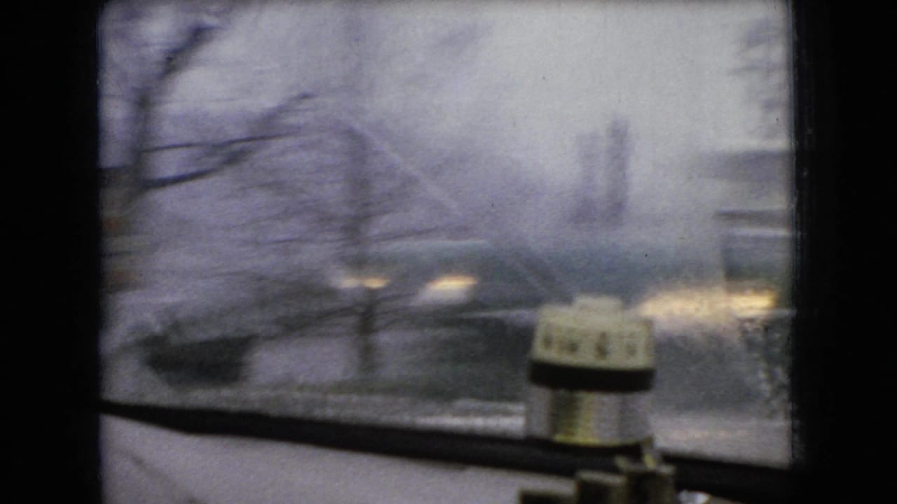 1966 Ohio 8mm home-movie frame showing a wet windshield view from inside a moving vehicle, with blurred roadway and a small cylindrical device on the dashboard.