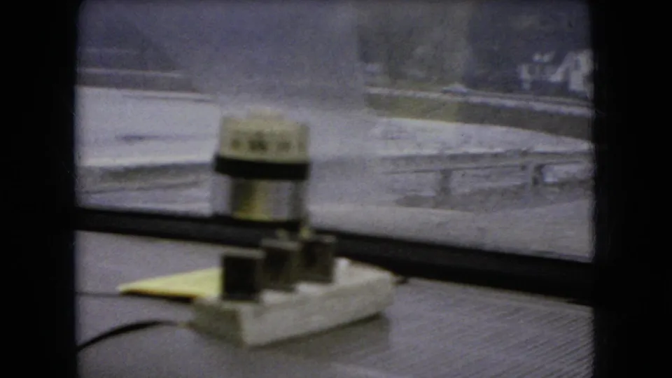 1966 Ohio 8mm frame looking past a small dashboard-mounted cylindrical device toward a rain-softened bridge railing and roadway outside the vehicle.
