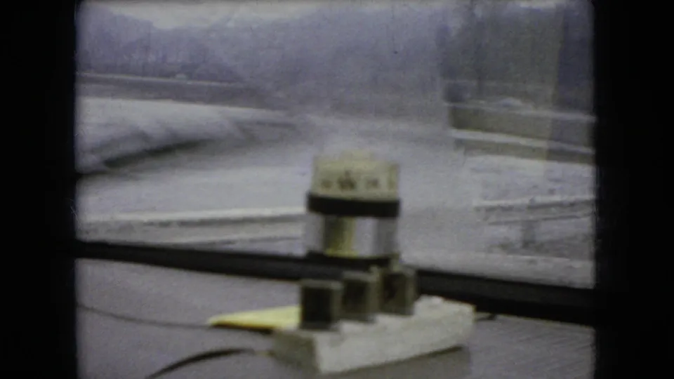 1966 Ohio 8mm frame from inside a moving car, looking across a rain-streaked windshield toward bridge railings and blurred trees with a small dashboard device in the foreground.