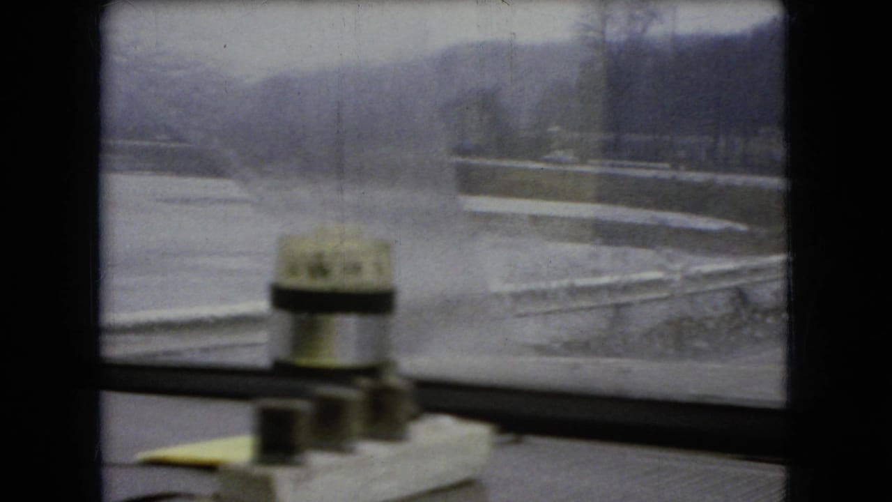 1966 Ohio 8mm frame looking through a rain-streaked car window toward water and a low roadside landscape, with a small windshield washer or wiper mechanism visible inside the vehicle.