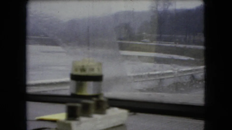 1966 Ohio 8mm frame looking through a rain-streaked car window toward water and a low roadside landscape, with a small windshield washer or wiper mechanism visible inside the vehicle.