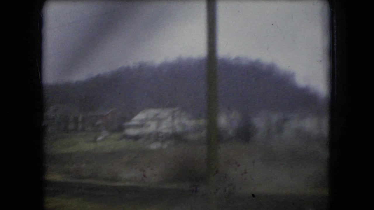 Blurred rainy landscape seen through a vehicle window with a wooded hill and pale roadside structure or groundform beyond.