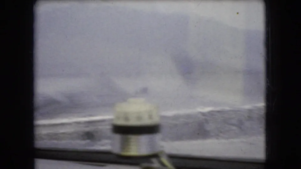 1966 Ohio 8mm frame looking past an interior windshield washer or wiper mechanism toward rain-blurred water, shoreline, and distant hills.