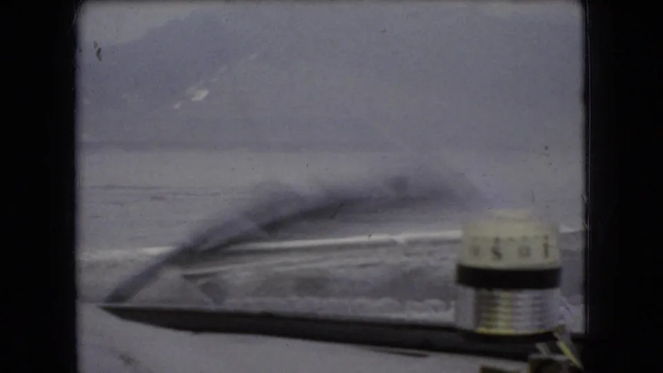 1966 8mm home-movie frame from supplied Ohio metadata, looking through a car windshield toward a gray water horizon with the wiper and hood visible.