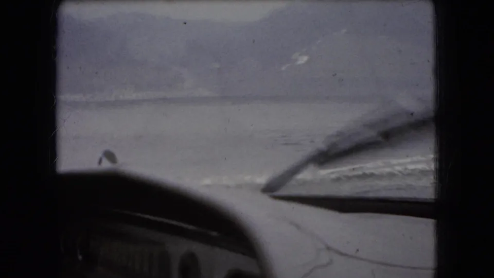 In-car 8mm frame with the rounded vehicle hood and windshield wiper silhouetted against a muted water horizon, with no identifiable person visible in this still.