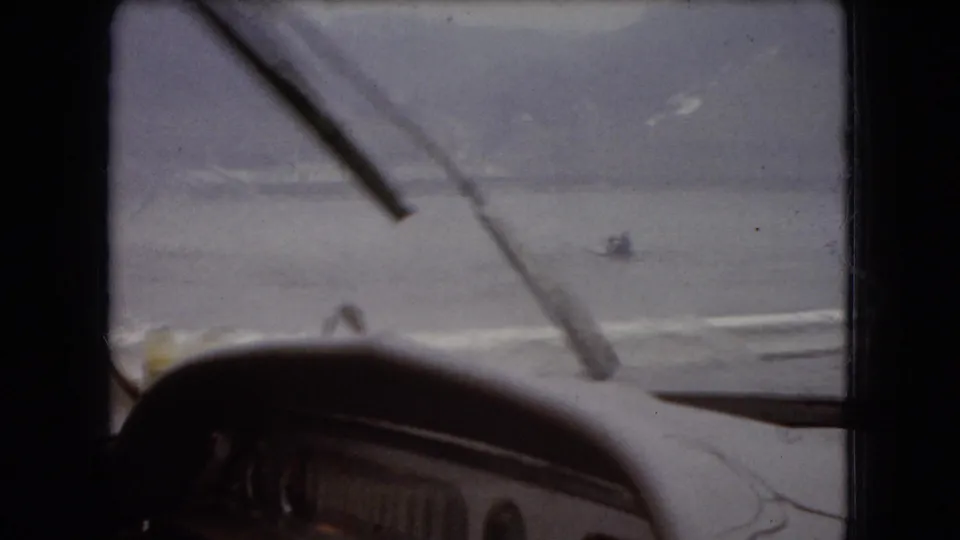 Passenger-side car-window view from a 1960s 8mm travel scene, showing wipers, wet glass, the vehicle hood, and a pale body of water beyond.