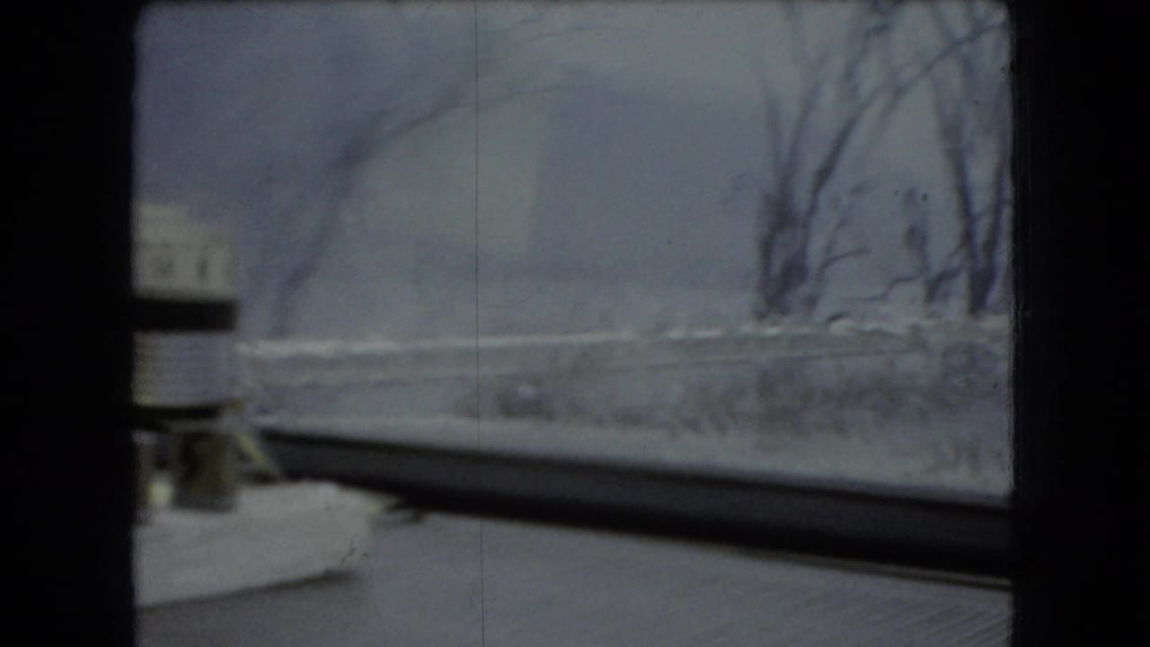 1966 Ohio 8mm frame showing a passenger-side view through the front windshield toward a blurred snowy roadside, bare trees, and a small white dashboard object.