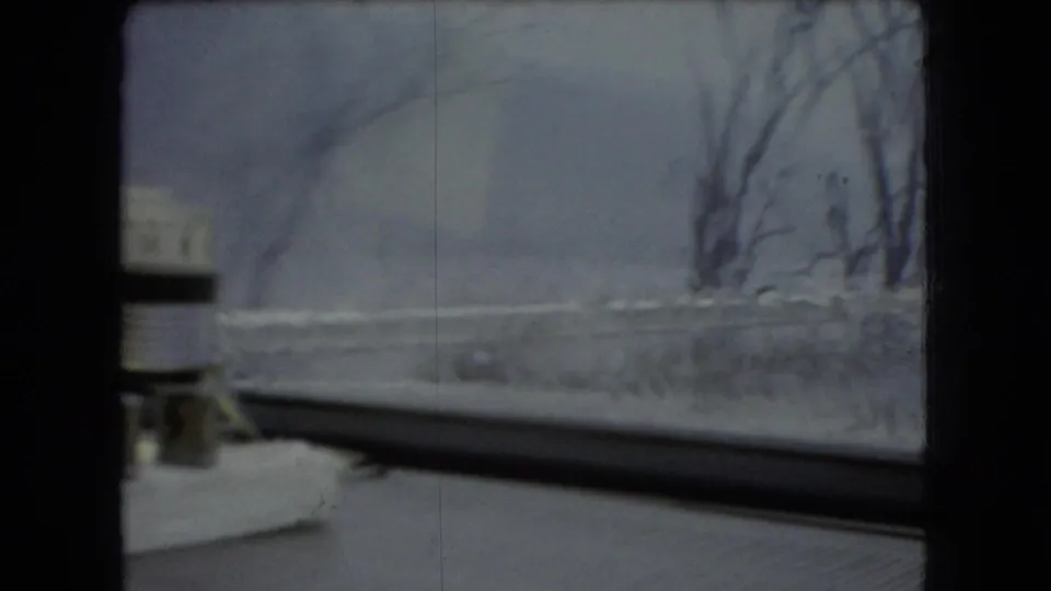 1966 Ohio 8mm frame showing a passenger-side view through the front windshield toward a blurred snowy roadside, bare trees, and a small white dashboard object.