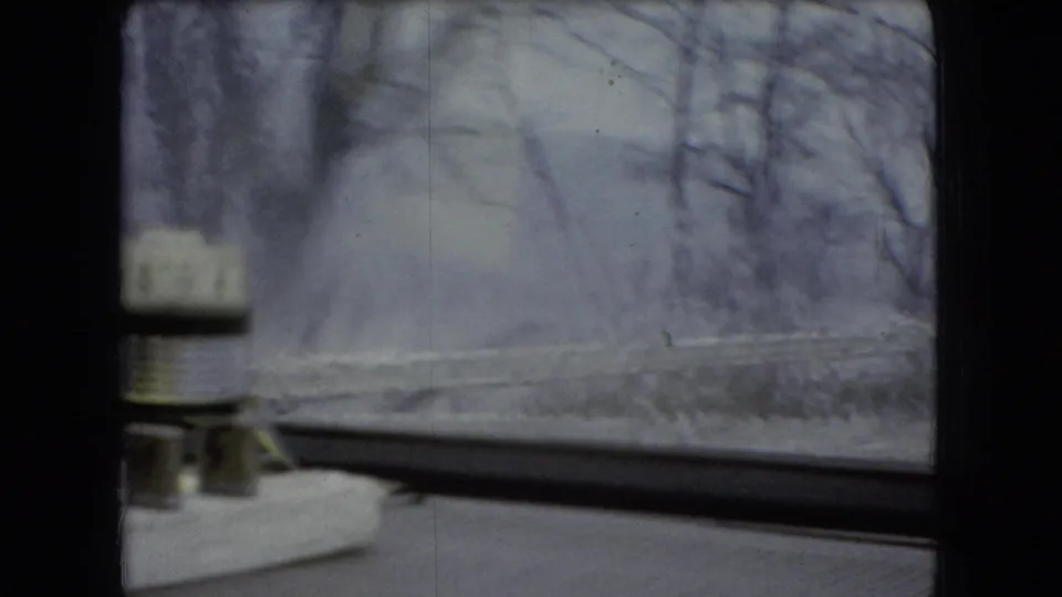 1966 Ohio home-movie frame from inside a moving car, looking past a dashboard-mounted object toward snow, bare trees, and a muted winter road edge.