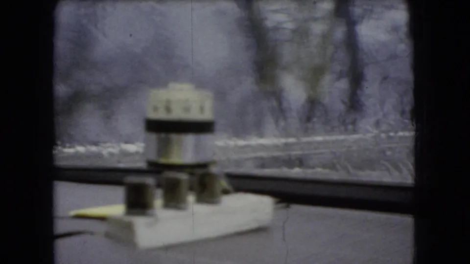 1966 Ohio 8mm frame showing the car interior framing a snowy roadside scene, with a white dashboard object prominent in the foreground and bare trees beyond the glass.