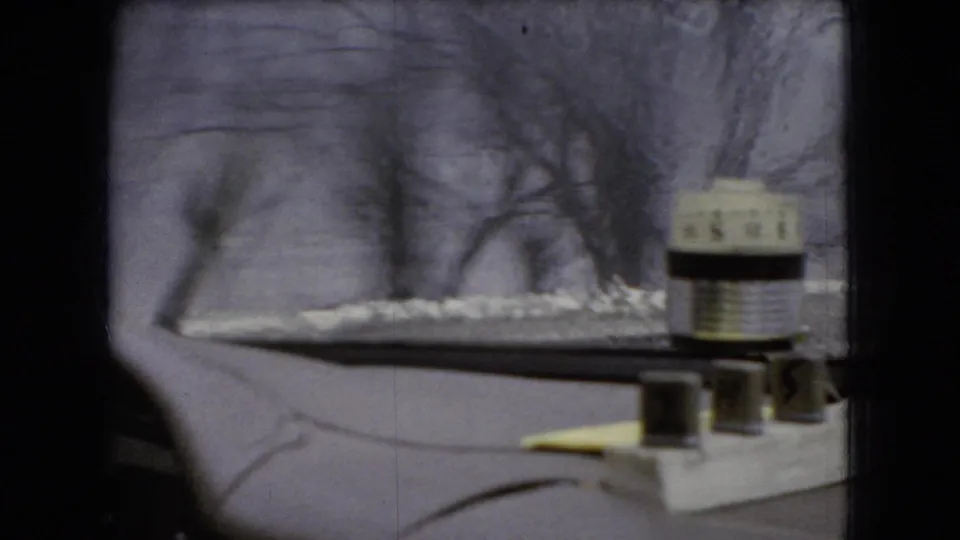 1966 Ohio home-movie frame looking through the windshield at a snowy road edge and bare trees, with the dashboard object and dark car interior visible in the foreground.