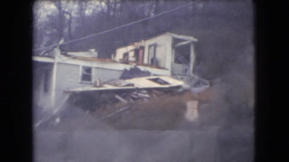 Rain-softened Ohio 1966 home-movie frame showing a low roadside house or structure with porch details, scattered materials, bare trees, and utility lines.