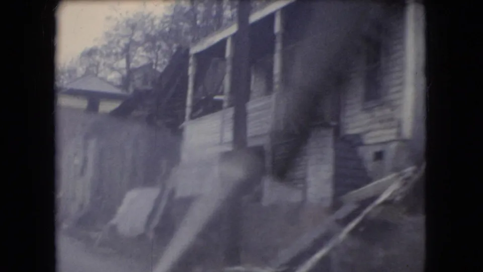 Close passing view from a rainy moving vehicle of a house porch, siding, windows, and yard edge in 1966 Ohio home-movie footage.