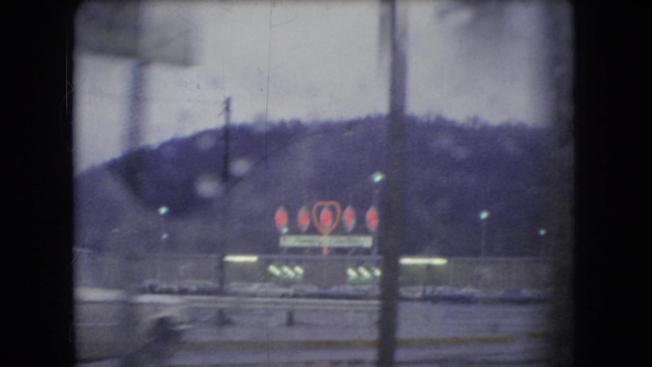 Lit roadside sign with red heart-like neon shapes below a wooded hillside.
