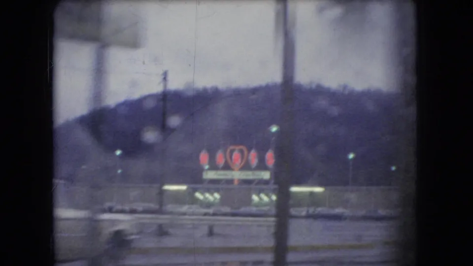 Distant roadside commercial sign with red heart-like neon shapes and small lights, seen against a dark Ohio hillside in 1960s home-movie footage.