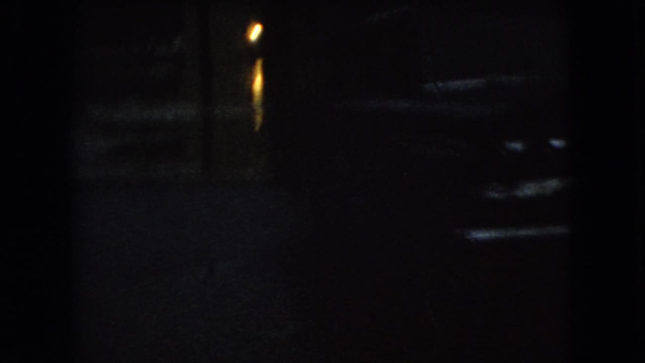 Dark 1966 Ohio 8mm frame showing a wet nighttime street, a parked car shape, and warm lamp reflections on the pavement.
