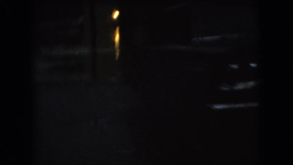 Dark 1966 Ohio 8mm frame showing a wet nighttime street, a parked car shape, and warm lamp reflections on the pavement.