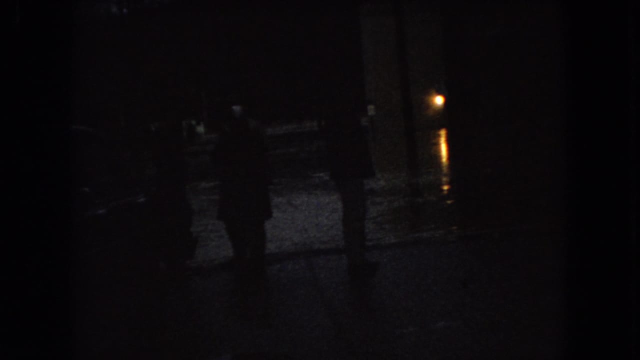 Low-light 1966 Ohio street view with silhouetted pedestrians, parked cars, and street lamp reflections on wet pavement.