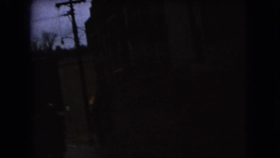 Underexposed 1966 Ohio 8mm frame with a pale sky patch, dark vertical structures, and indistinct street-side foreground forms.
