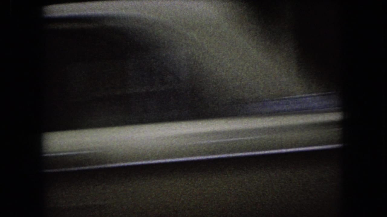 Dark 1966 Ohio home-movie frame with a blurred roadside or building facade and a pale horizontal street edge under low light.