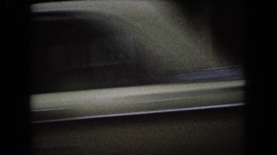 Dark 1966 Ohio home-movie frame with a blurred roadside or building facade and a pale horizontal street edge under low light.