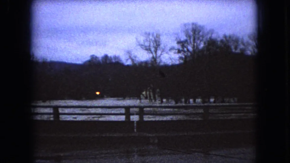 1966 Ohio 8mm frame of a snowy roadside at dusk with bare winter trees, horizontal fence or guardrail lines, and a small distant headlight or lamp.