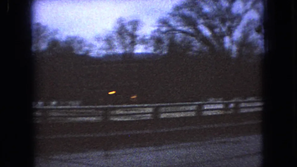 1966 Ohio 8mm frame showing glowing car lights near a snowy roadside fence or guardrail with bare trees silhouetted against a cold evening sky.