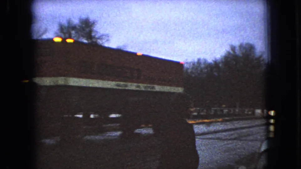 1966 Ohio 8mm frame showing a large dark roadside structure or passing vehicle with small lights, snow along the ground, and bare trees in the background.