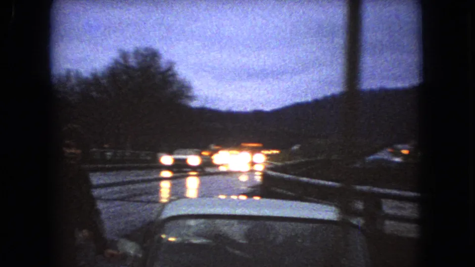 1966 Ohio 8mm frame from inside a moving vehicle, showing a wet road at dusk with oncoming headlights reflecting on pavement, guardrails, wooded hills, and the car hood at the lower edge.