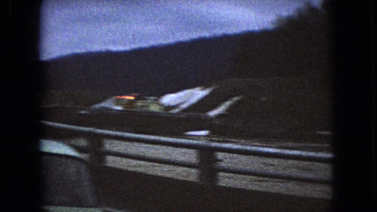 Blurred roadside view with guardrail, dark hillside, wet pavement, and a passing vehicle shape.